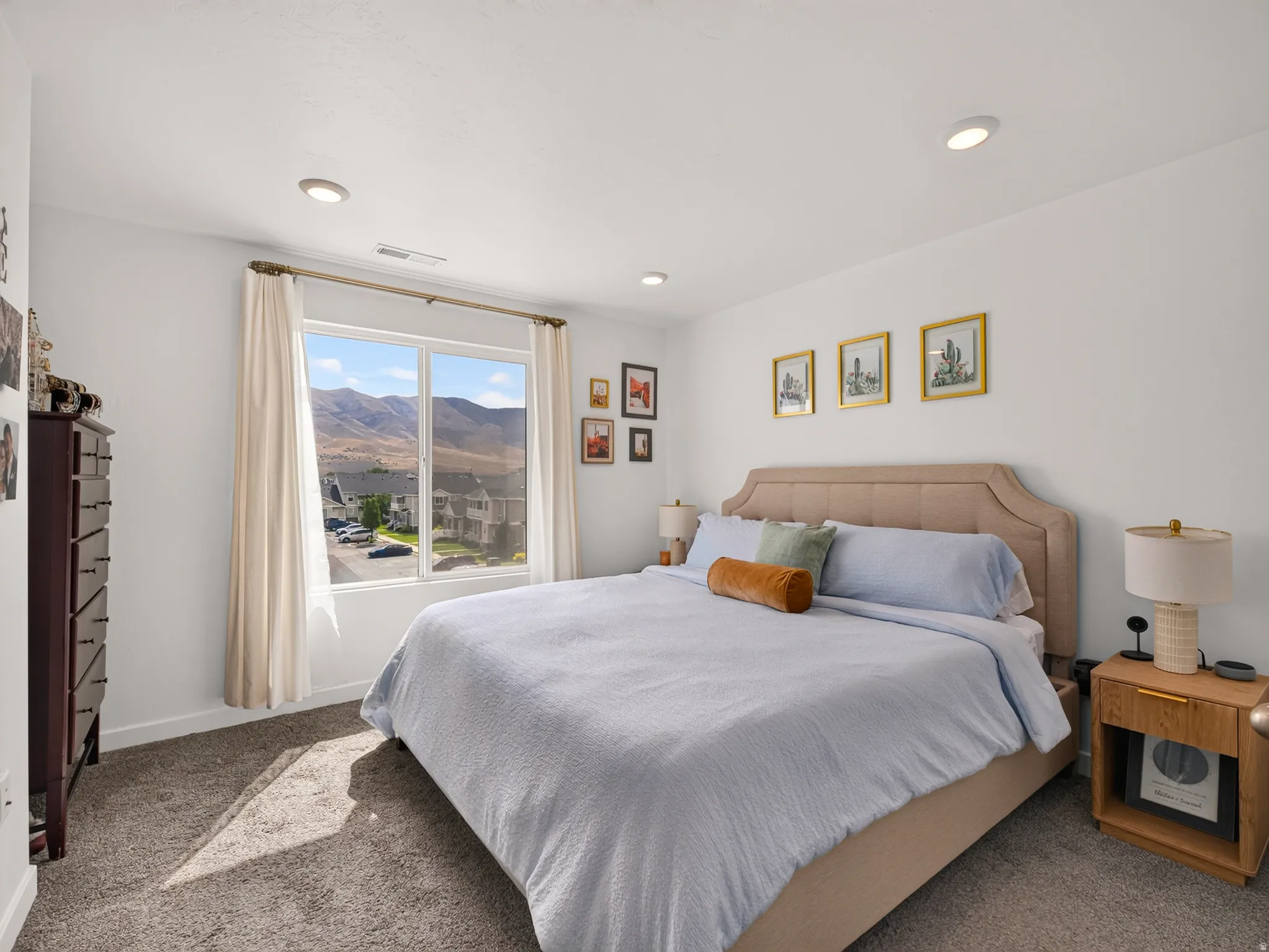 Bright, light-filled bedroom featuring expansive windows framing mountain scenery, neutral wall finishes, recessed ceiling lighting, wood-finish nightstands, and carpet flooring throughout
