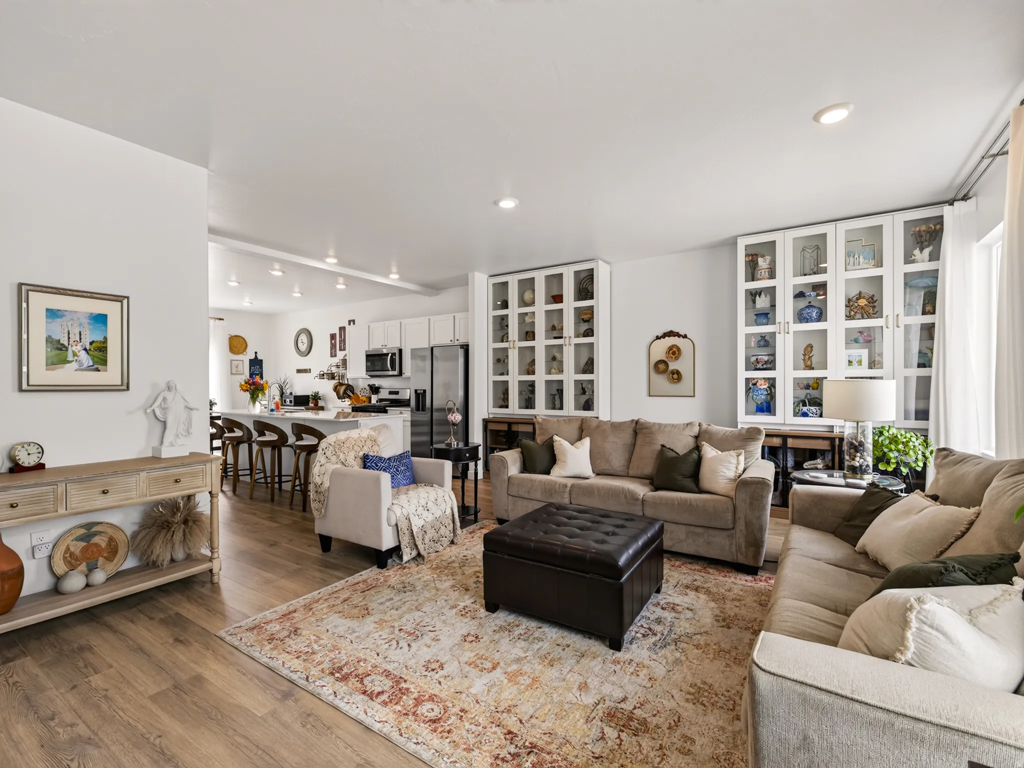 Wood-finish flooring throughout, integrated recessed lighting, white built-in cabinetry with glass panel doors, a kitchen island with seating, and stainless steel appliances