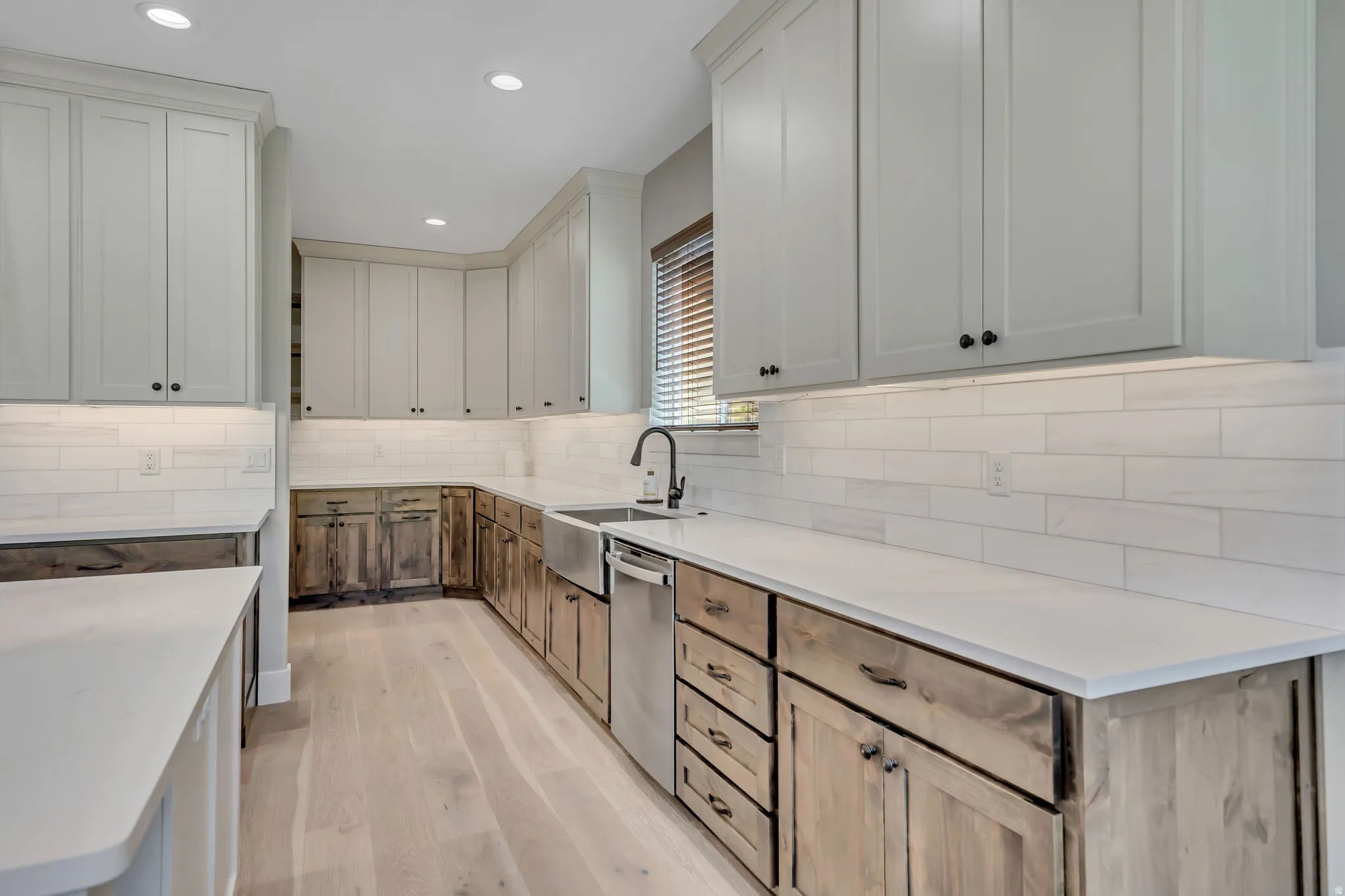 Recessed lighting illuminating a spacious kitchen with light wood-finish flooring, white subway tile backsplash, white countertops, and a stainless steel farmhouse sink