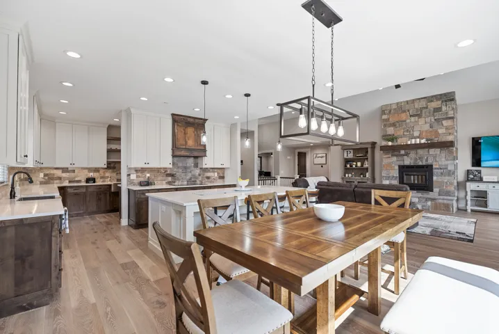 Open-concept living area featuring wood-finish flooring, a stone-clad fireplace with a wood mantle, a kitchen island with seating, white cabinetry, and a wood range hood