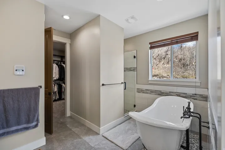 Freestanding soaking tub with dark-finish hardware, glass-enclosed shower, tiled flooring, and a window with wood-finish blinds
