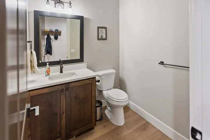 Hardwood-style flooring, a dark wood-finish vanity, white countertop, black-framed mirror, and a three-light vanity fixture