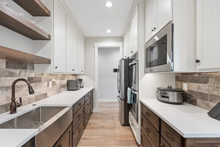 Galley kitchen with white shaker cabinetry, wood-finish base cabinets, light-toned countertops, and a tile backsplash