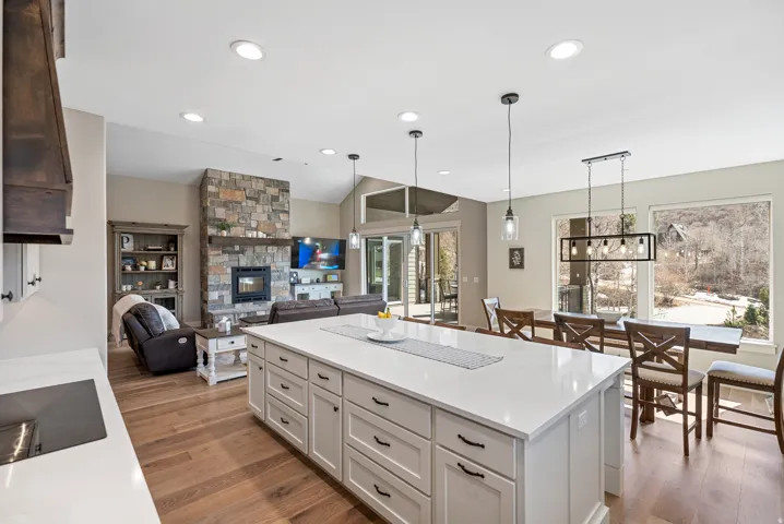 Open-concept living space with wood-finish flooring, a stone-faced fireplace, a large kitchen island with light-toned countertops, and expansive windows
