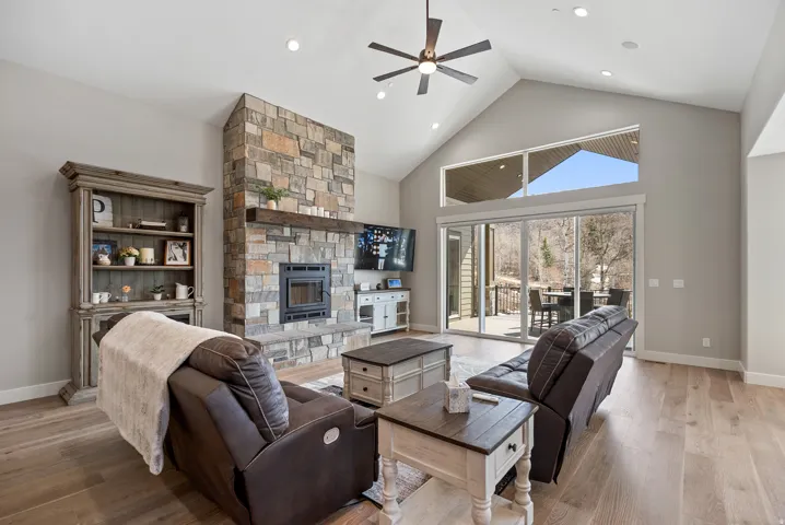 Vaulted ceiling with a ceiling fan, stone-clad fireplace with a hearth, wood-finish flooring, and expansive glass sliding doors