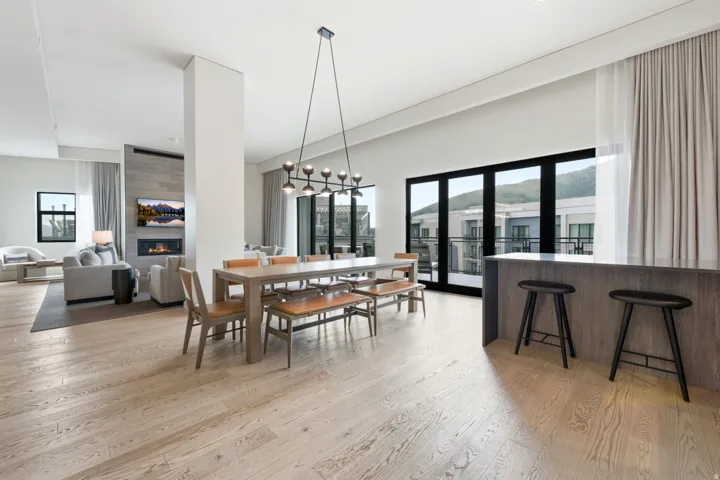 Light wood-finish flooring throughout, modern linear chandelier, built-in fireplace with a stone-finish facade, large black-framed glass doors, and a built-in bar with a countertop
