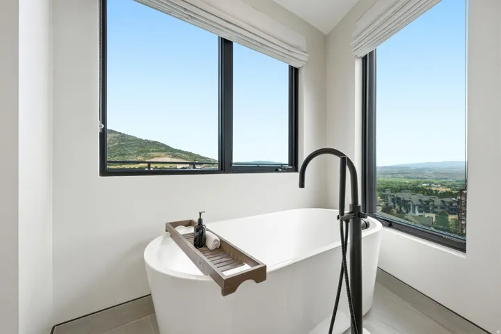 Freestanding soaking tub with a matte black floor-mounted faucet, large windows, and neutral-toned flooring