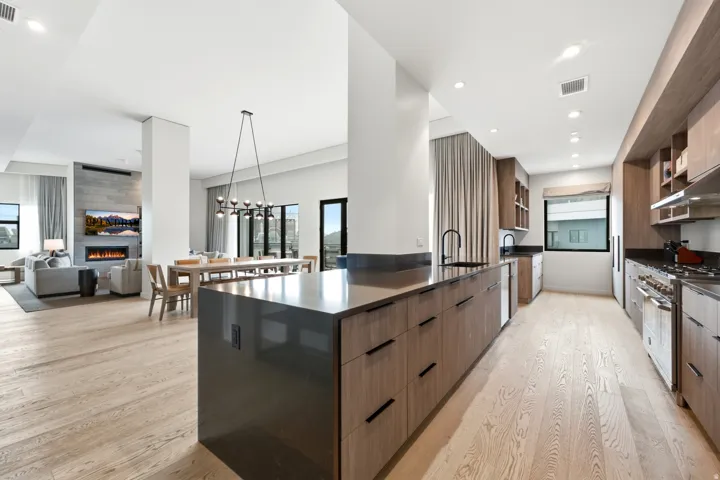 Open-concept living space with light wood-finish flooring, a contemporary kitchen featuring integrated appliances, a substantial kitchen island with dark countertops, and a living area centered around a stone-faced fireplace