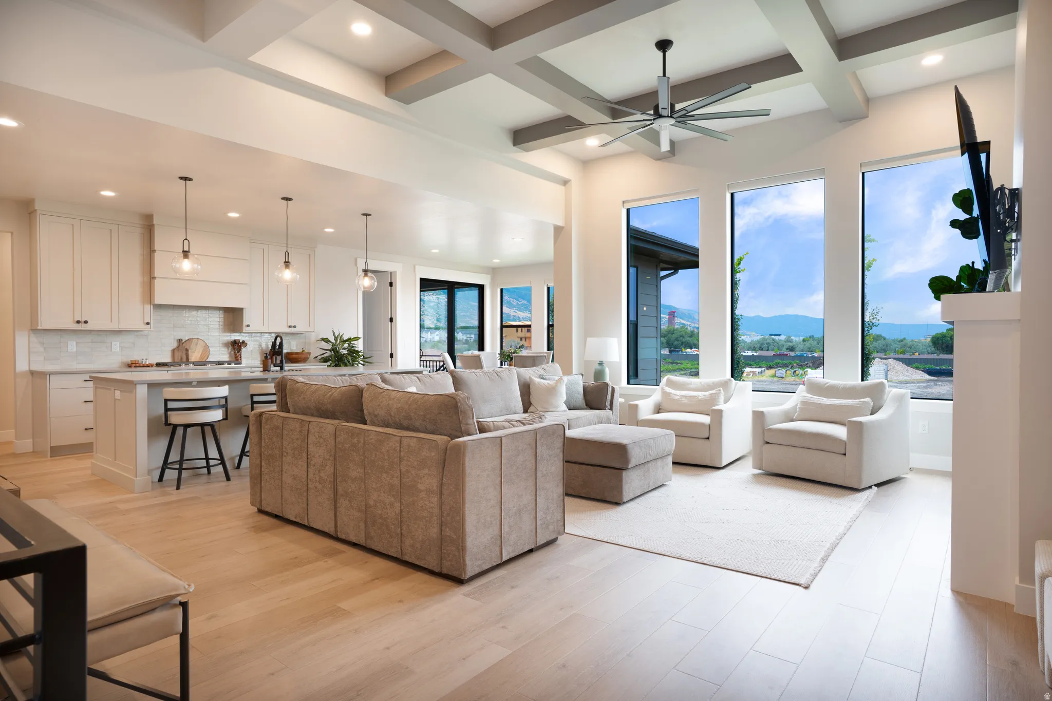 Open-concept living space with light wood-finish flooring, a coffered ceiling with recessed lighting, and expansive windows
