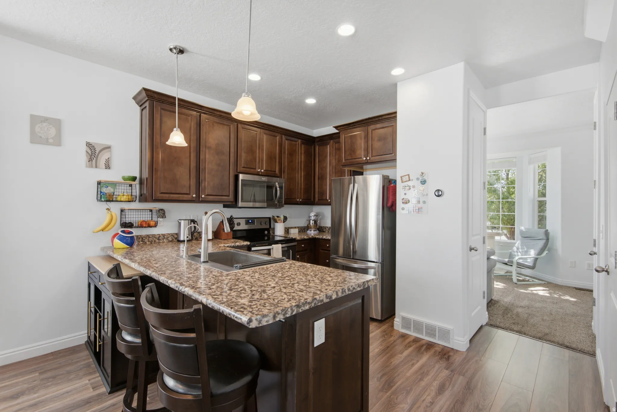 Kitchen featuring wood-finish flooring, dark wood cabinetry, a granite-style countertop island with an integrated sink, stainless steel appliances, and recessed lighting