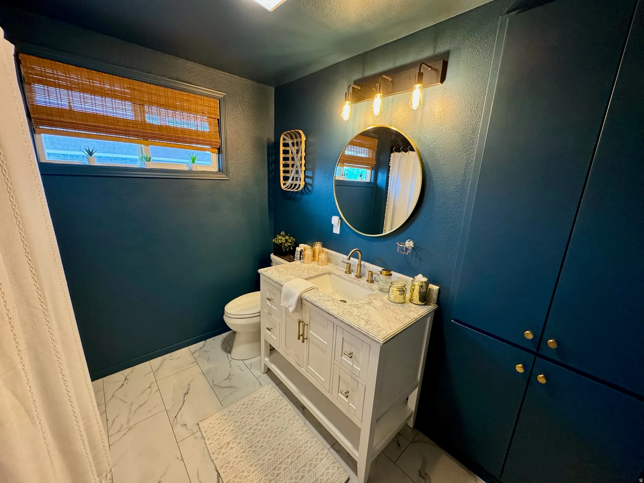 White vanity with a light-toned countertop, a gold-framed circular mirror, and built-in vertical cabinetry with gold-toned hardware