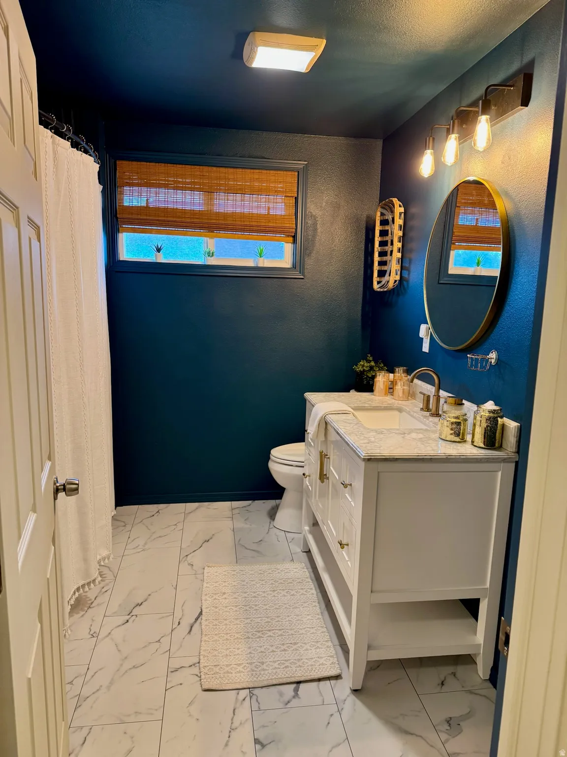 White vanity with a stone countertop, modern wall-mounted light fixture, oval mirror, and large format tile flooring