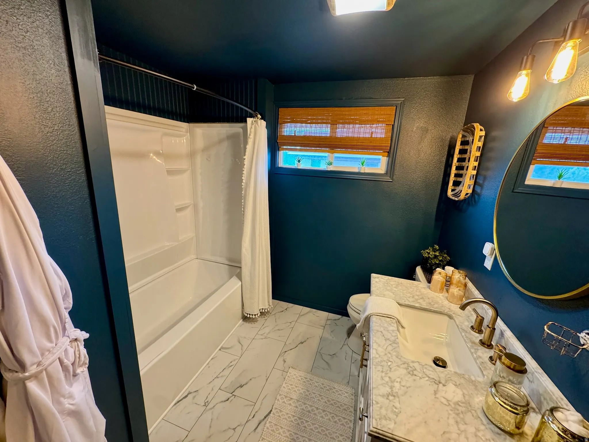 White tub/shower combination, dark blue walls, rectangular windows with blinds, white vanity with integrated sink, and gold-finish faucet