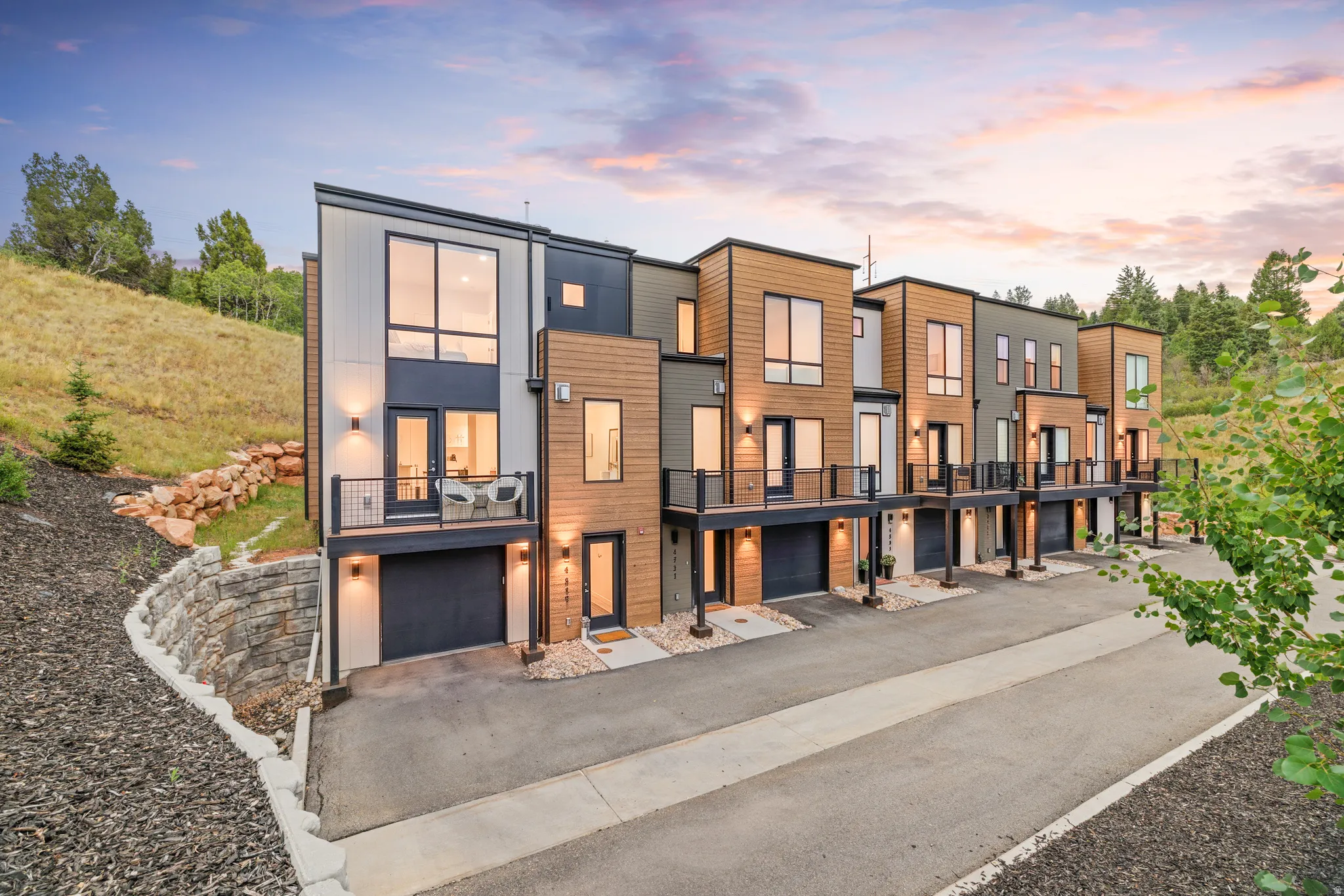 Contemporary townhome exteriors featuring wood-look and vertical panel siding, individual balconies with black railings, integrated single-car garages, and modern exterior lighting