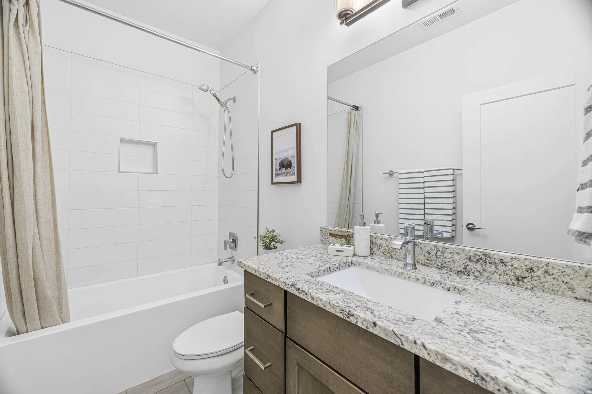 White tiled tub and shower combination with an integrated niche, a single vanity featuring a countertop with natural stone finish, and a large wall-mounted mirror
