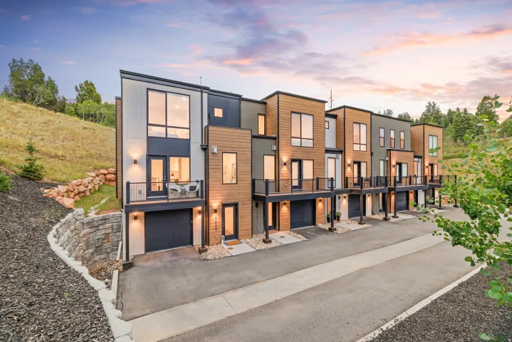 Contemporary townhome exteriors featuring wood-look and vertical panel siding, individual balconies with black railings, integrated single-car garages, and modern exterior lighting