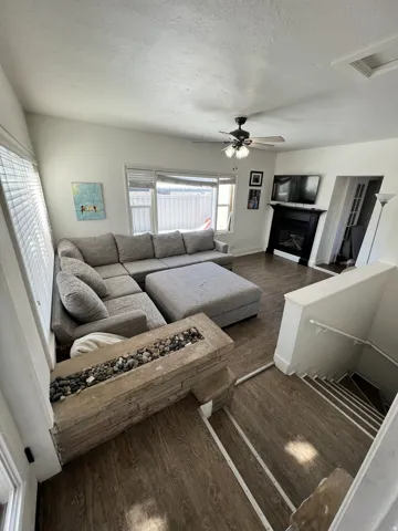 Living area featuring wood-finish flooring, a built-in fireplace with a dark mantle, a ceiling fan with integrated lighting, and a built-in stone fire pit