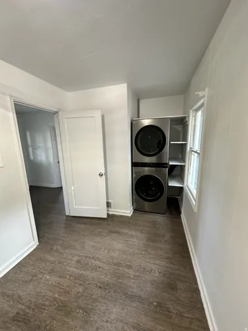 Stacked stainless steel laundry appliances, wood-finish flooring, white paneled walls, and built-in shelving