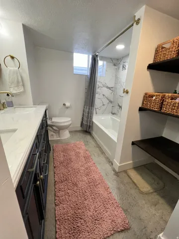 Vanity with dual sinks and gold-finish fixtures, built-in shelving, shower-over-tub with marble-look tile surround, and concrete-finish flooring