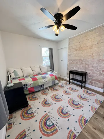 Exposed brick accent wall, wood-finish flooring, ceiling fan with integrated lighting, built-in storage cabinet with double doors, and a window with white trim