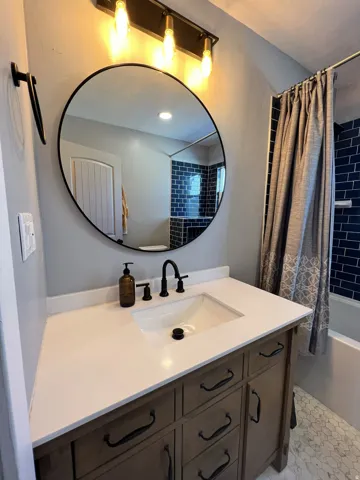 Wood-finish vanity with an integrated sink and matte black faucet, complemented by a round mirror and overhead lighting