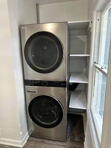Stacked stainless steel laundry appliances, built-in shelving, wood-finish flooring, and a large window