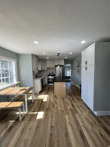 Open-concept kitchen featuring wood-finish flooring, recessed lighting, a central island with a dark countertop, stainless steel appliances, and white cabinetry
