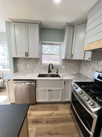 White shaker-style cabinetry, herringbone tile backsplash, stainless steel farmhouse sink with a black faucet, wood-finish flooring, and a stainless steel appliance suite