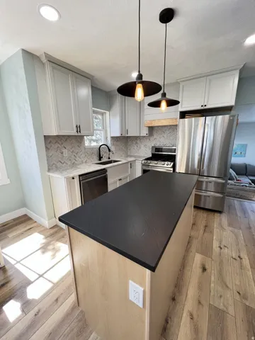 Kitchen featuring a central island with a dark countertop, light wood-finish flooring, white cabinetry, stainless steel appliances, and a herringbone pattern tile backsplash