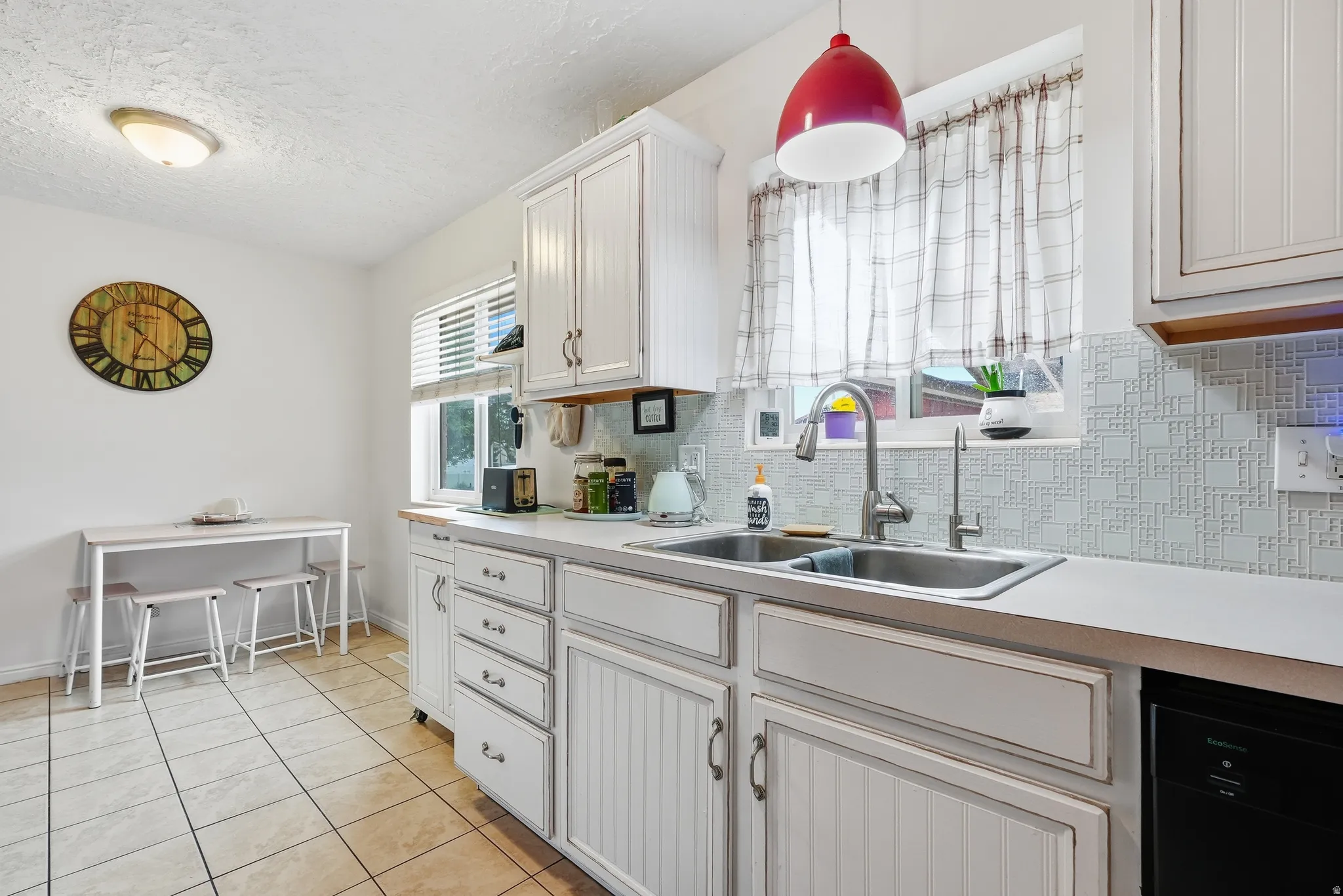 White cabinetry, light-toned countertops, stainless steel double-basin sink, patterned tile backsplash, and ceramic tile flooring
