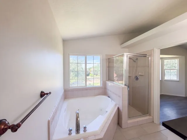 Corner soaking tub with tiled surround, glass-enclosed shower, dual windows, and tiled flooring
