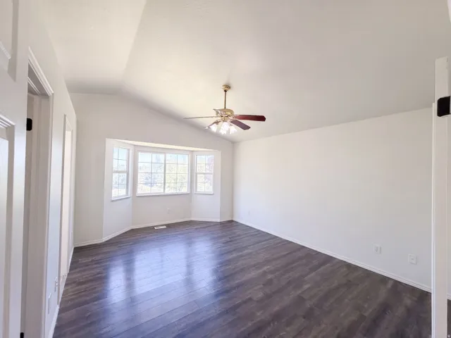 Vaulted ceiling, bay window, wood-finish flooring, ceiling fan with light, and white walls