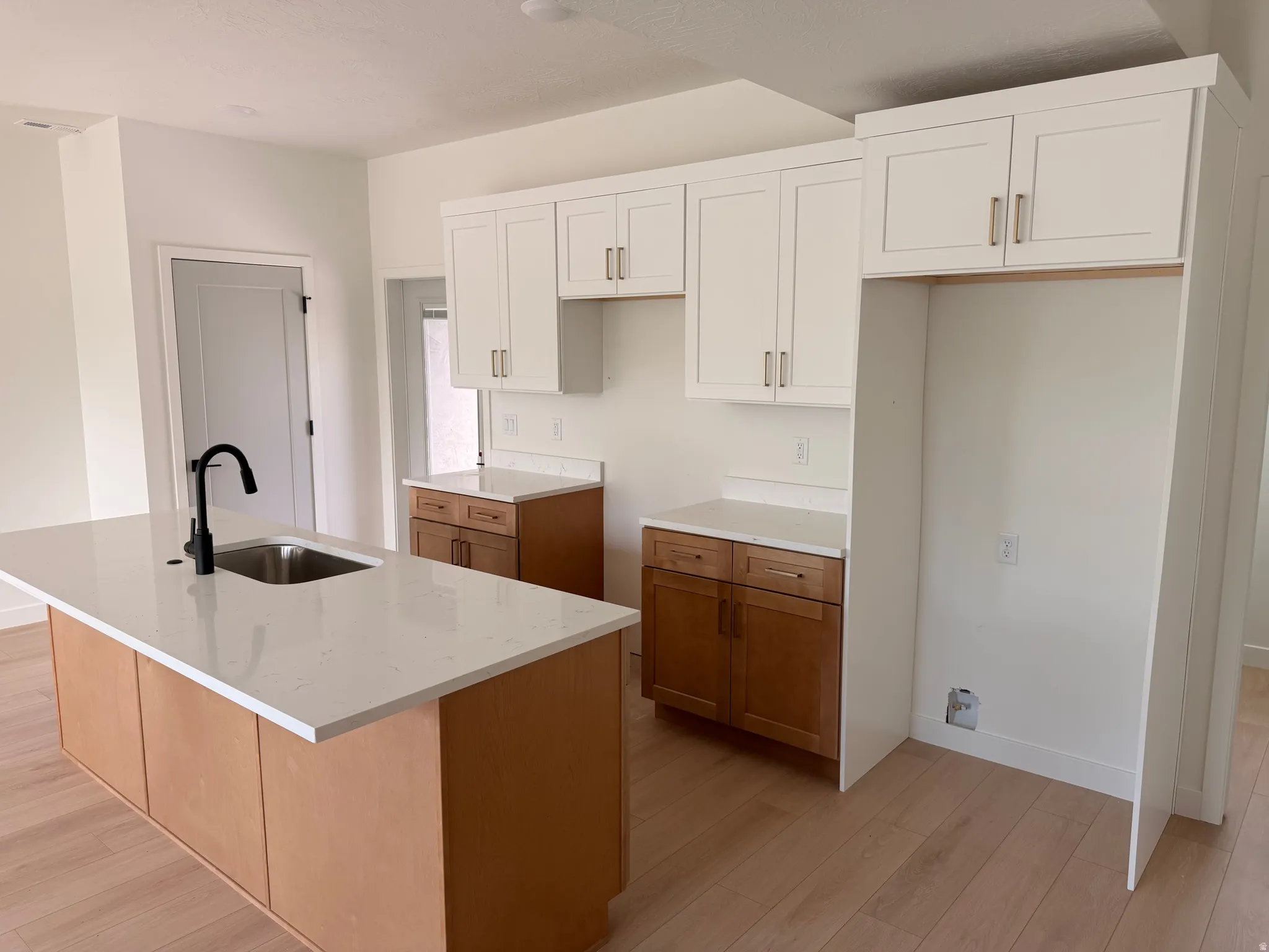 Kitchen featuring a central island with an undermount sink and matte black faucet, light wood-finish flooring, white shaker-style upper cabinetry, and wood-finish lower cabinetry with white countertops
