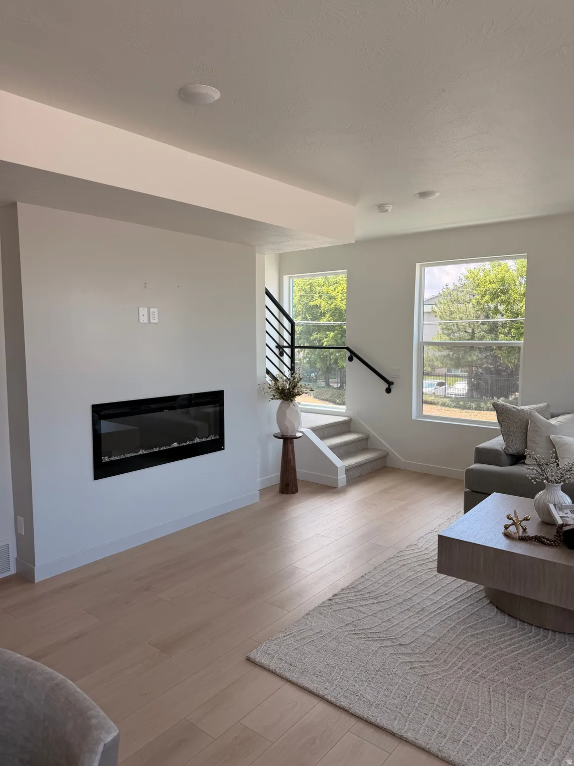 Recessed electric fireplace, light wood-finish flooring, two large windows, modern black metal stair railing, and recessed ceiling lighting