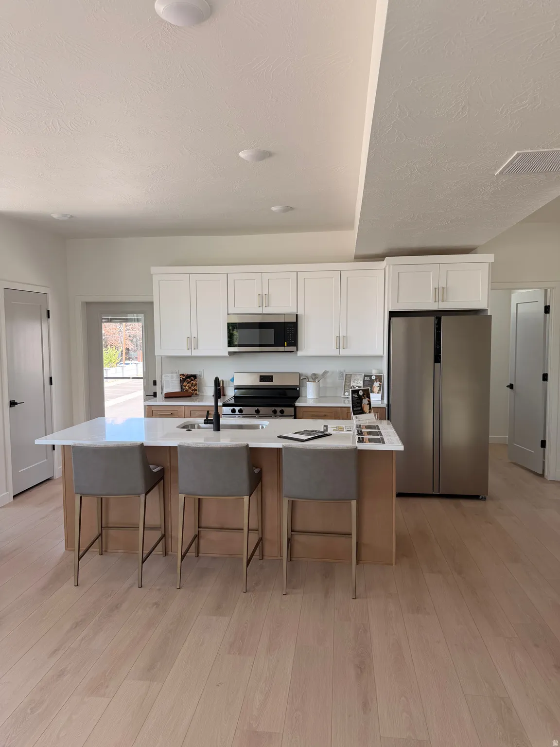 Central kitchen island with a white countertop and seating for three, complemented by stainless steel appliances, white cabinetry, and wood-finish flooring