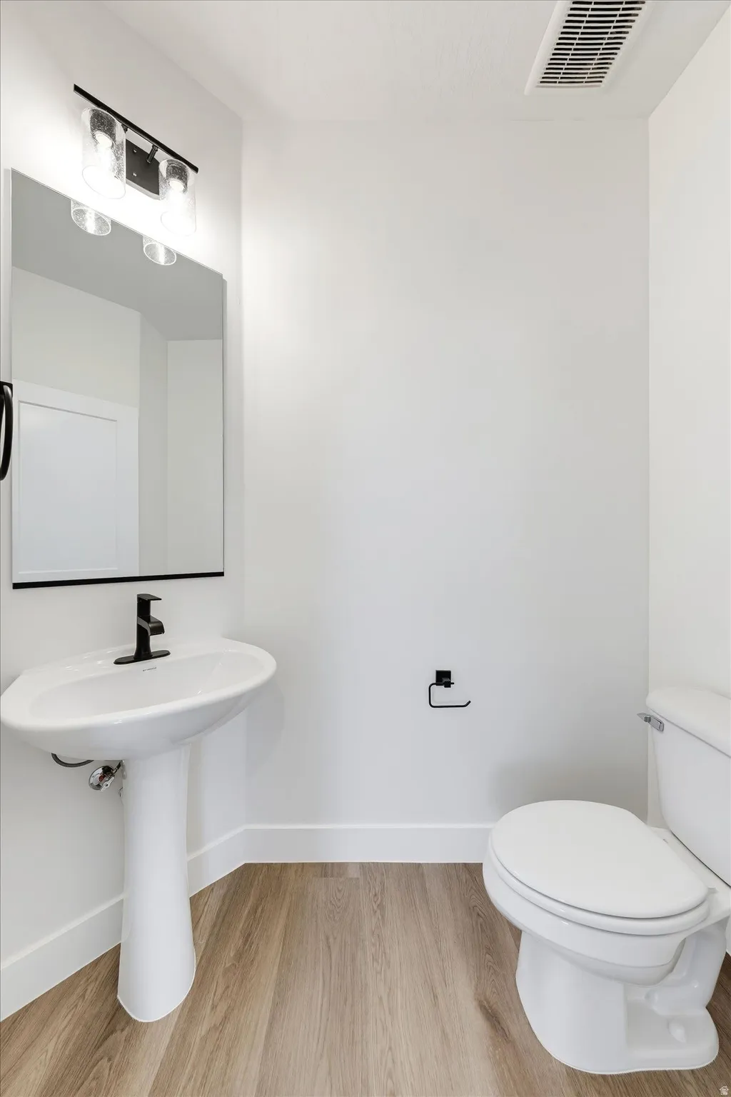 Pedestal sink with a matte black faucet, rectangular mirror, contemporary vanity light fixture, and wood-finish flooring