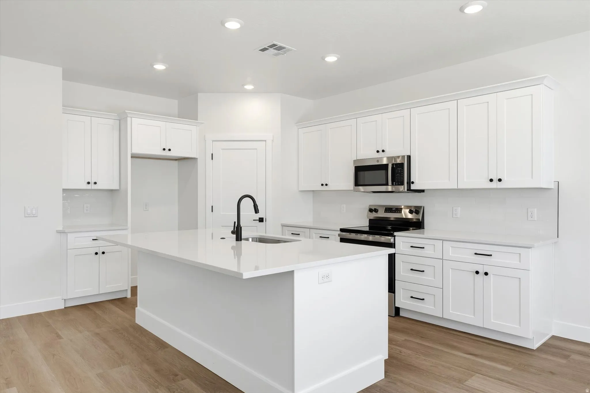 Kitchen featuring white shaker cabinetry, stainless steel appliances, a center island with sink, quartz countertops, and wood-finish flooring