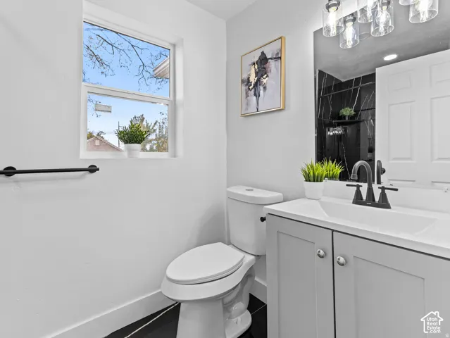 White single-pane window, black wall-mounted towel bar, white toilet, gray vanity with white countertop and integrated sink, matte black faucet, and clear glass vanity lighting