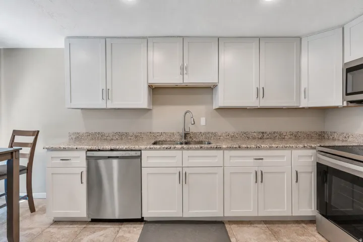 White shaker-style cabinetry, stainless steel dishwasher, undermount sink with a gooseneck faucet, light-toned stone-patterned countertops, and tile flooring