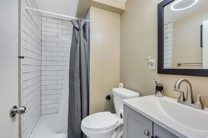 White subway tile shower surround, single vanity with light gray cabinetry, integrated sink, brushed nickel faucet, and a wall-mounted mirror with a dark frame