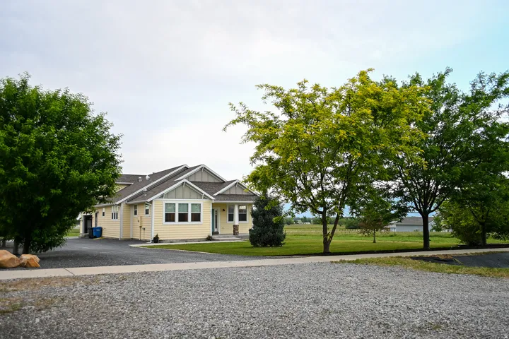 Single-story home featuring light yellow siding, a covered front porch, a gravel driveway, and a manicured lawn with mature trees