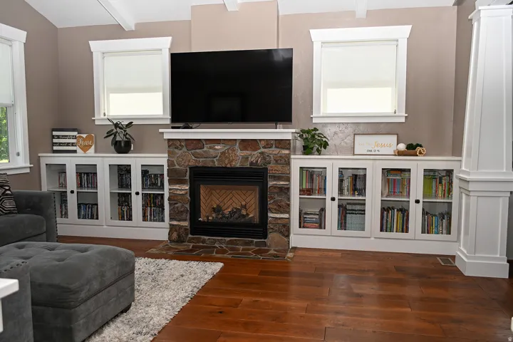 Stone-clad fireplace with hearth, wood-finish flooring, two large windows, white built-in cabinetry with glass doors, and exposed ceiling beams