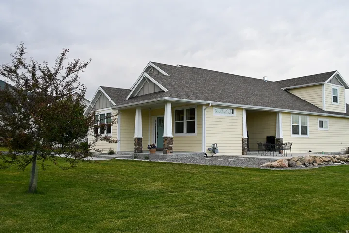 Expansive lawn, covered front porch with stone-clad columns, covered back patio, dormer window, and light-colored siding
