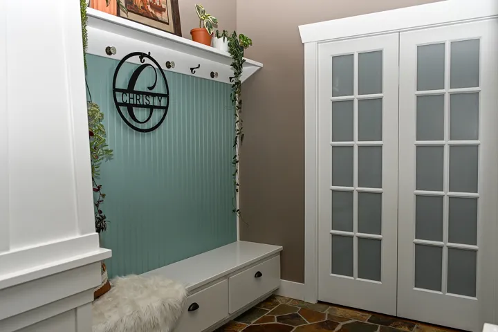 Built-in bench with storage, beadboard wall paneling, upper shelf with coat hooks, French doors with frosted glass, and natural stone flooring