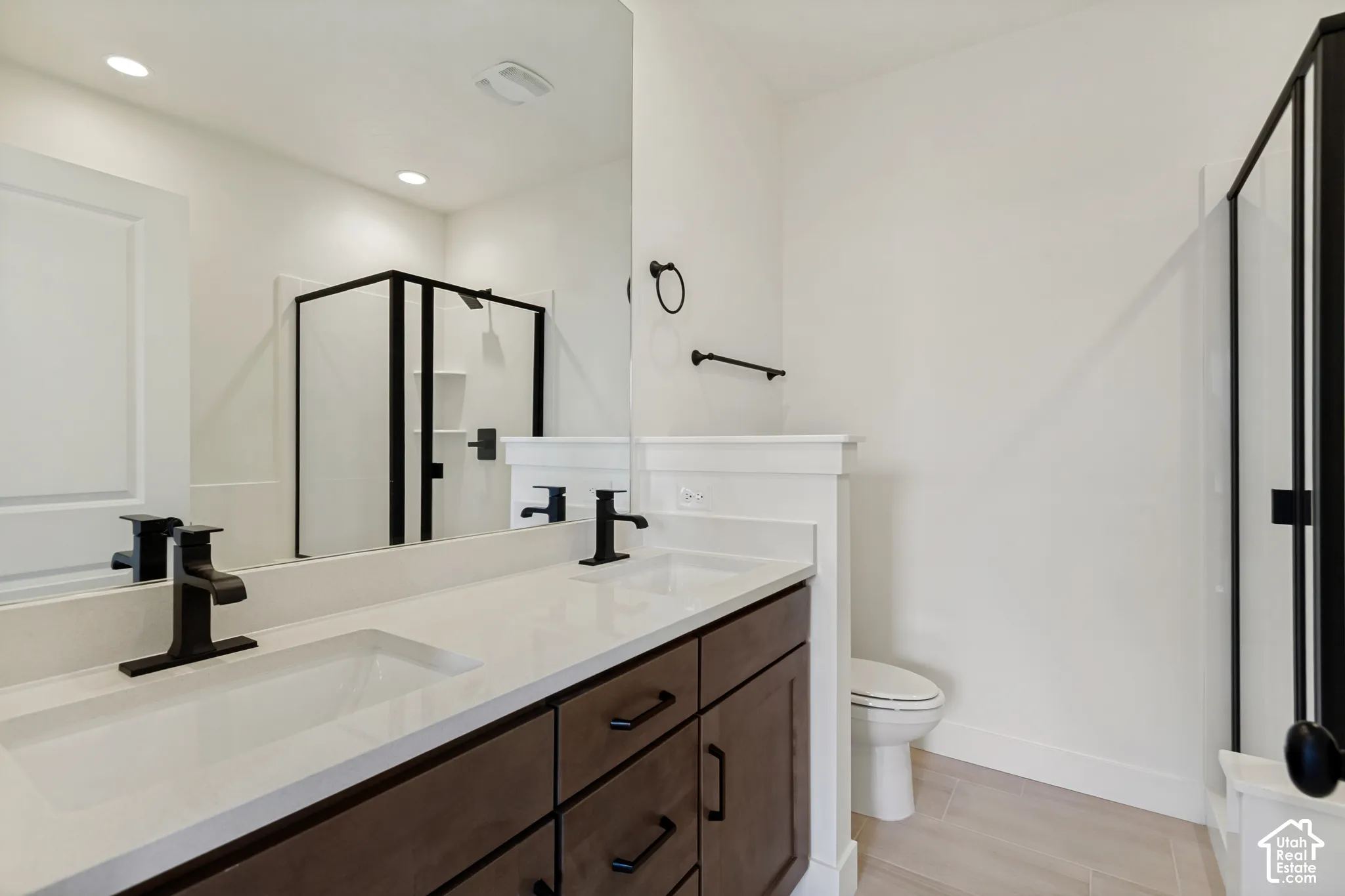 Dual vanity with matte black fixtures, integrated sinks, white countertop, dark wood-finish cabinetry, and a glass-enclosed shower with black framing