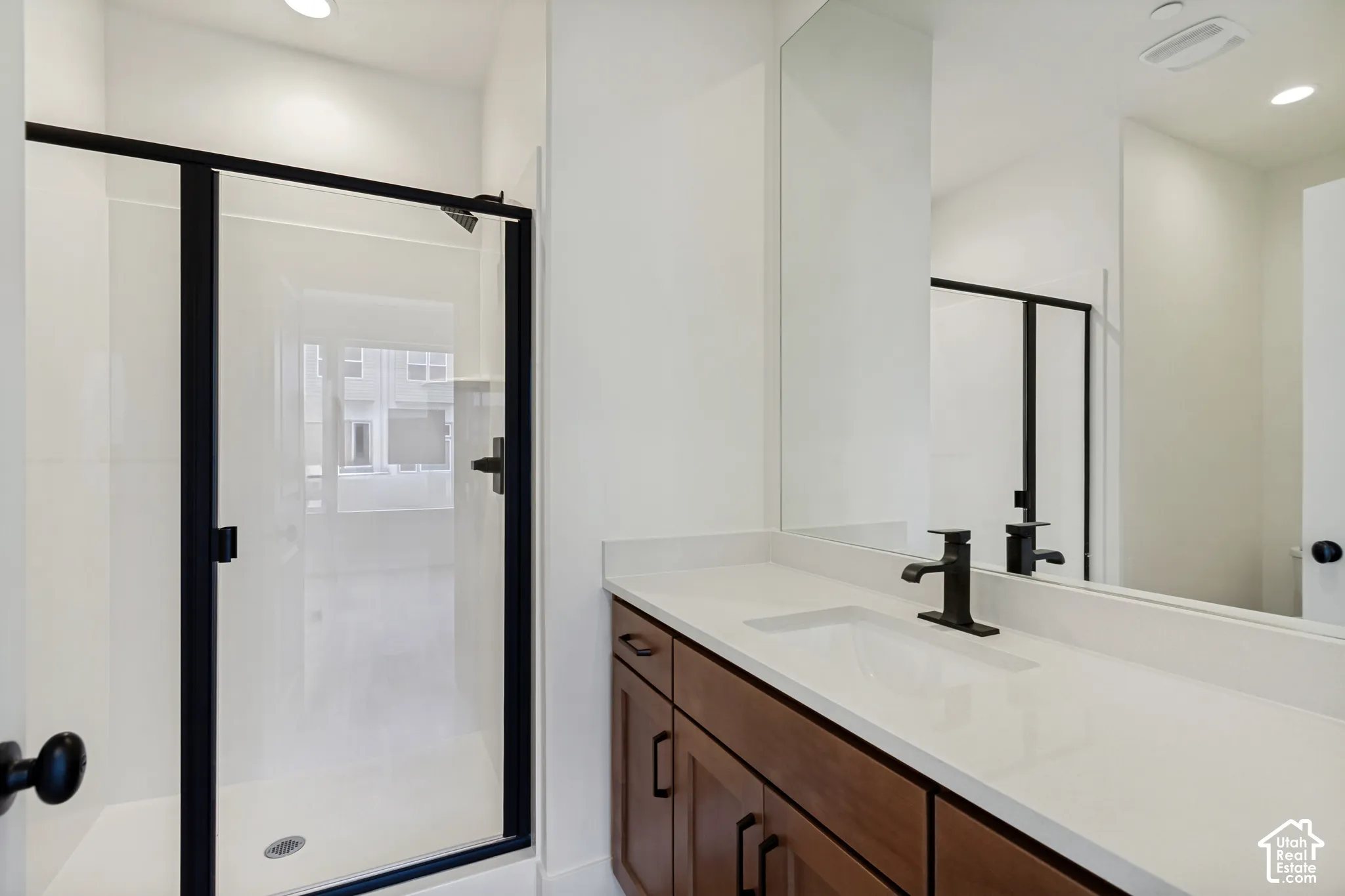 Walk-in shower with black-framed glass enclosure, single vanity with wood cabinetry, light-toned countertop, and undermount sink, complemented by a matte black faucet