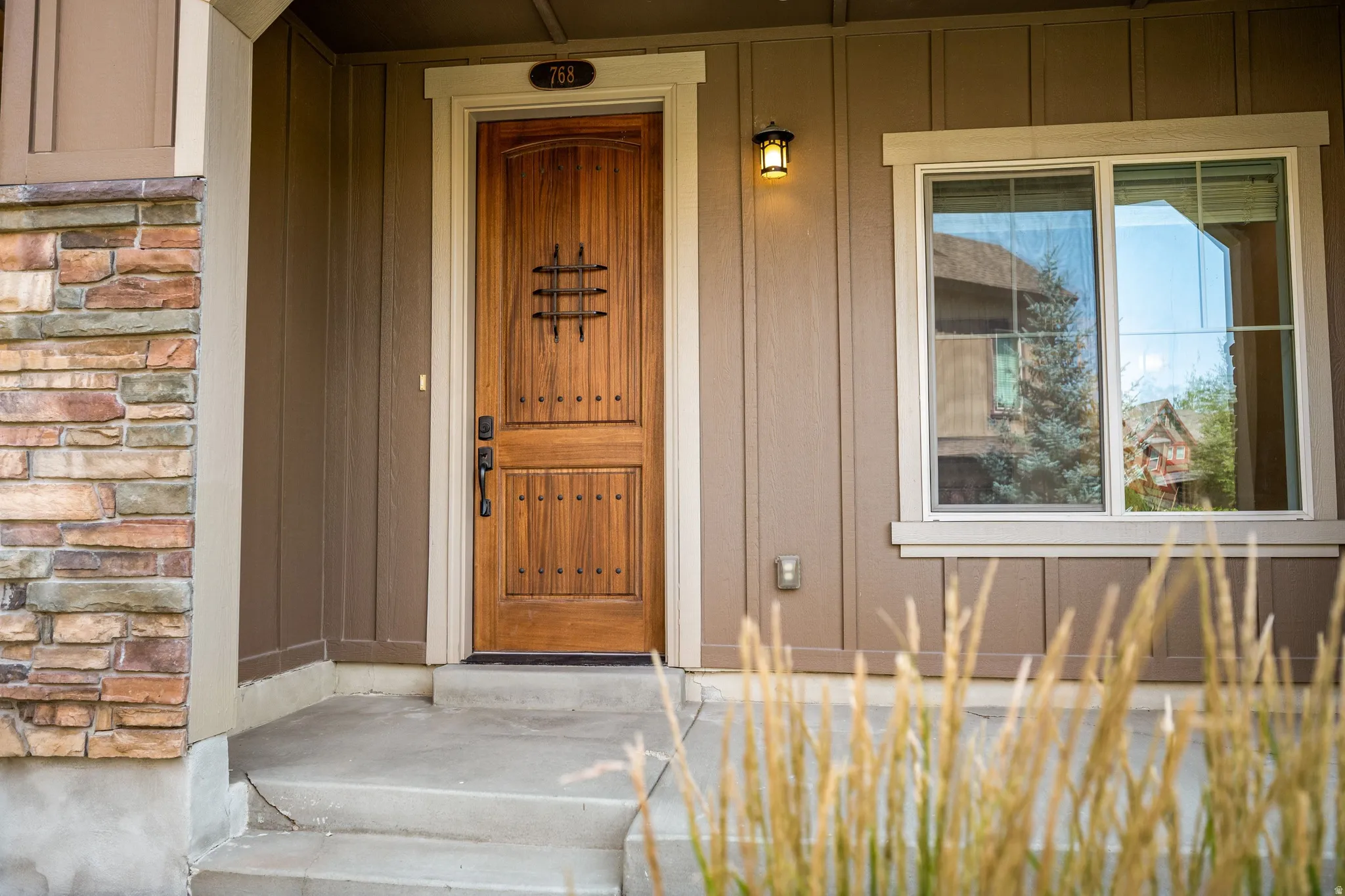 Crafted wood-paneled door with decorative ironwork, stone veneer column, concrete porch, and exterior lighting fixture