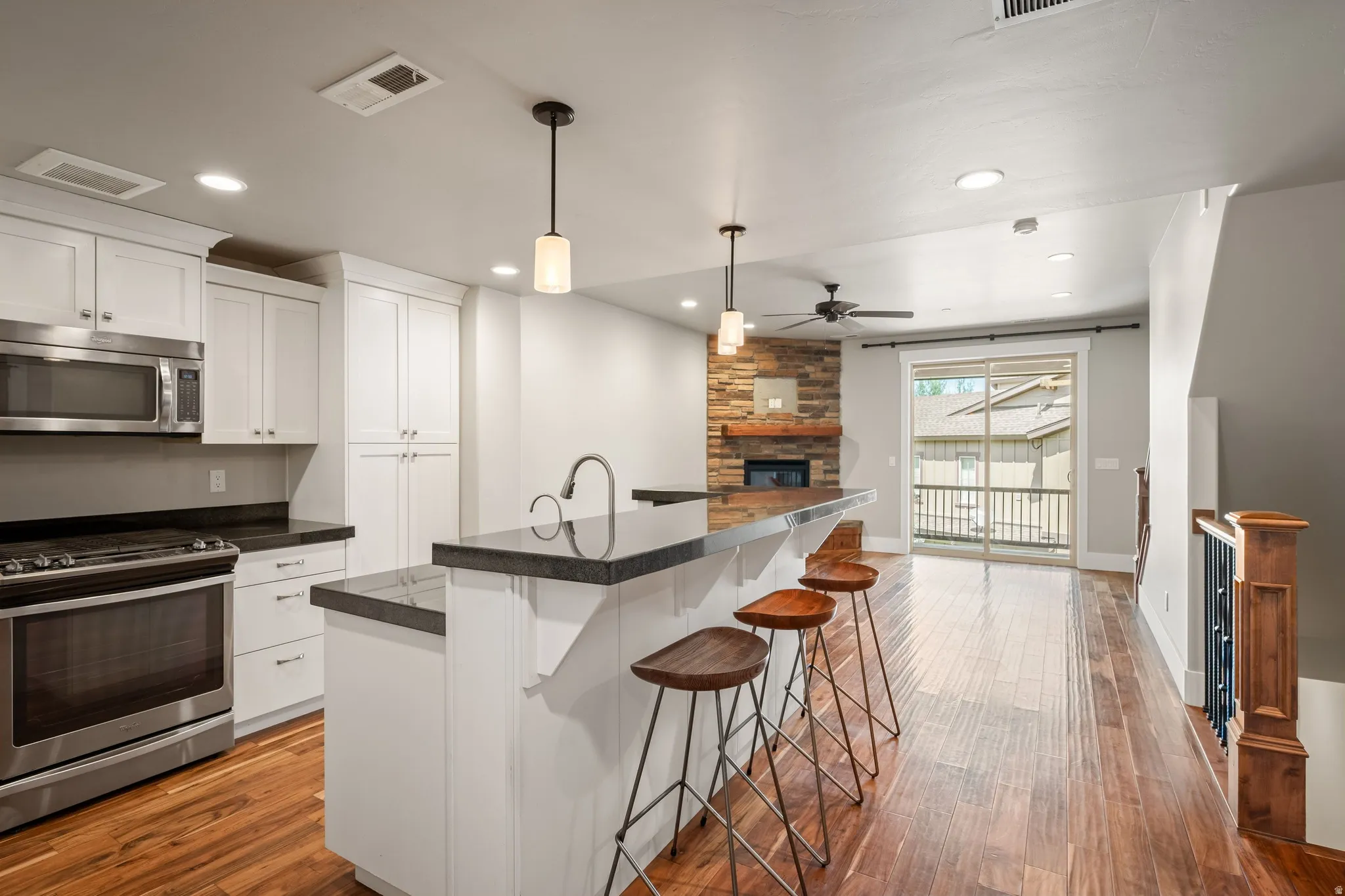 Open concept living area featuring wood-finish flooring, a kitchen island with dark countertops, white cabinetry, stainless steel appliances, and a fireplace with a stone surround