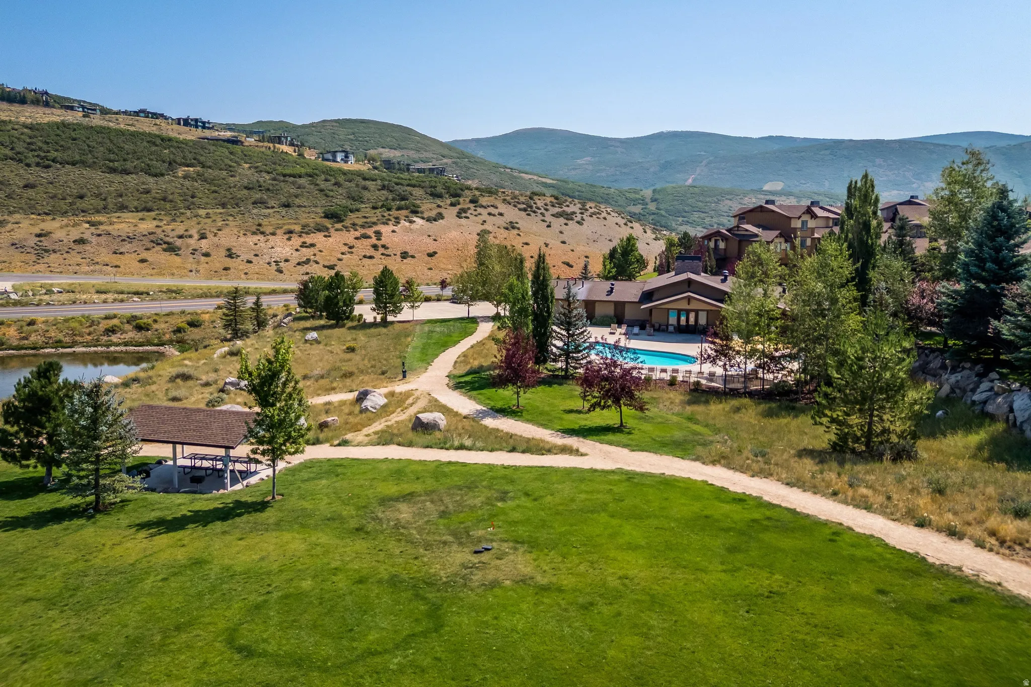 Expansive green lawn with a winding path, featuring a covered picnic area, a lake, and a swimming pool with surrounding lounge chairs