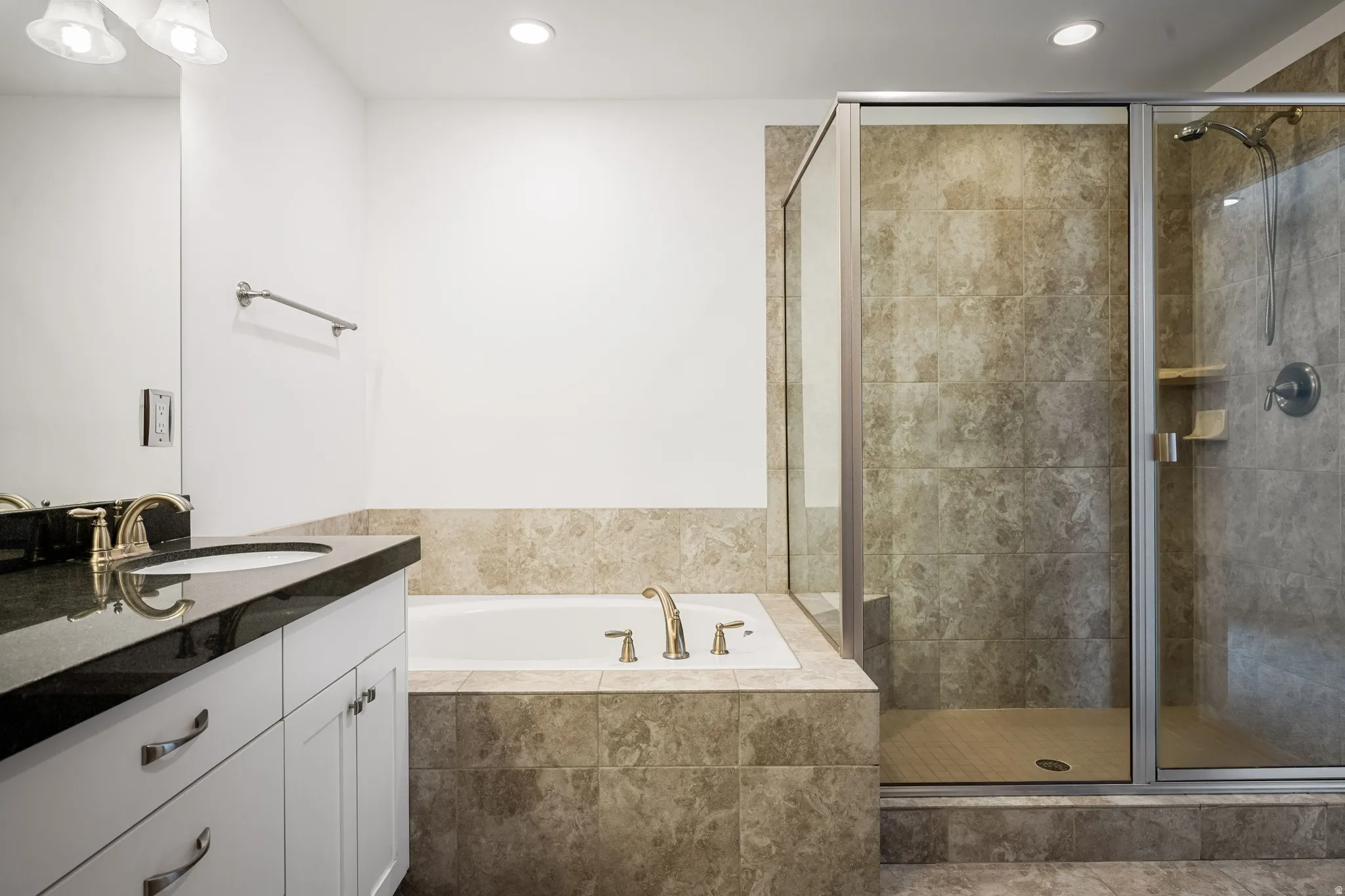 Vanity with a dark countertop and undermount sink, built-in bathtub, and a glass-enclosed shower with tiled walls